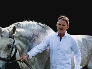 Man in white shirt leading a tall flea-bitten grey horse in a bridle