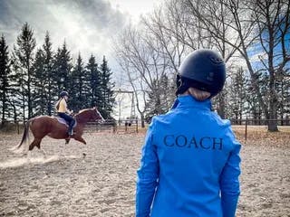 Coach in a red jacket standing in an arena giving a lesson to a young lady on a chestnut horse.