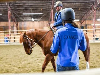 Coach with a blue jacket teaching a male riding western and wearing a helmet