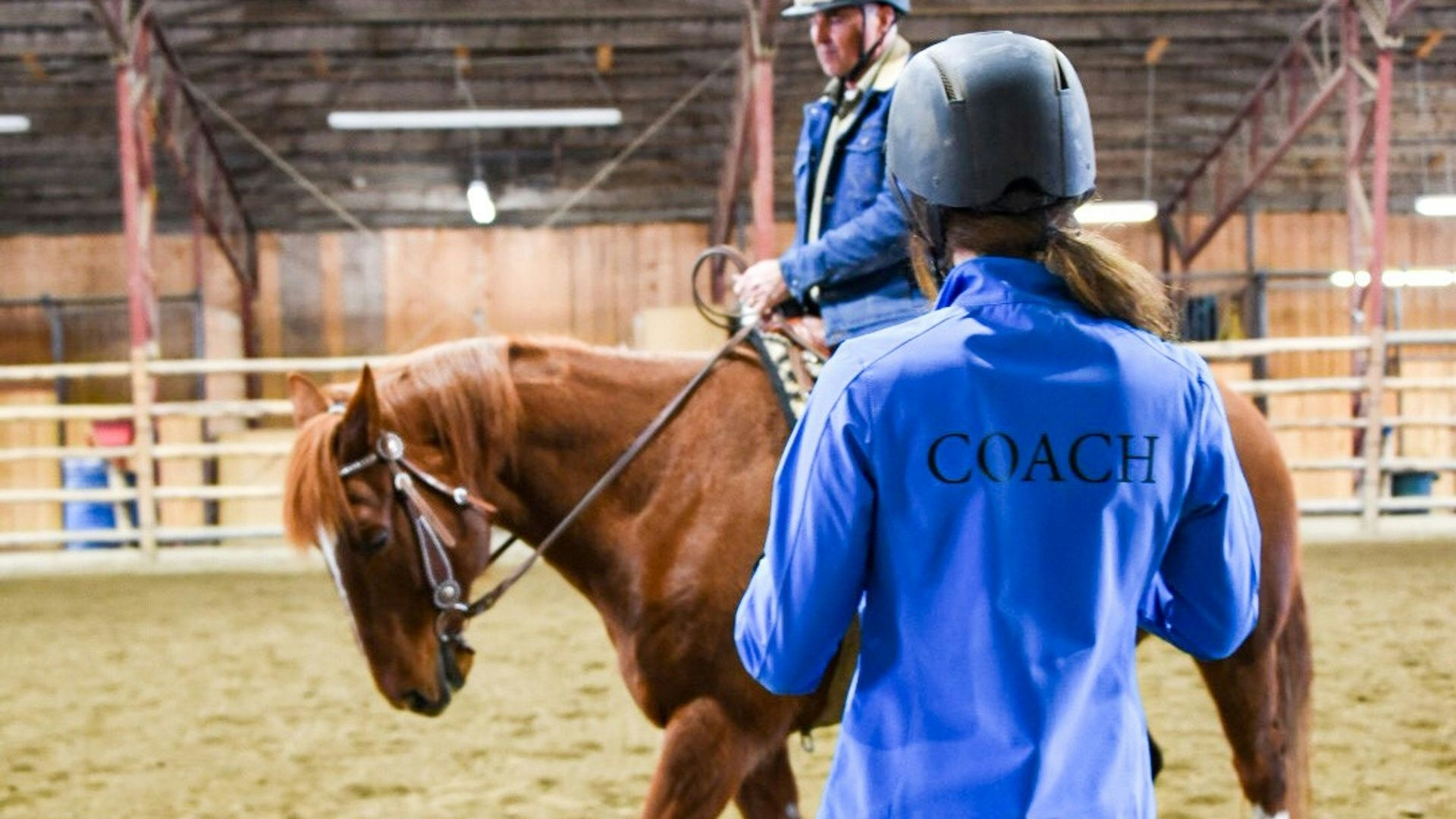 An instructor stands in the foreground as a mature male student rides a chestnut horse.