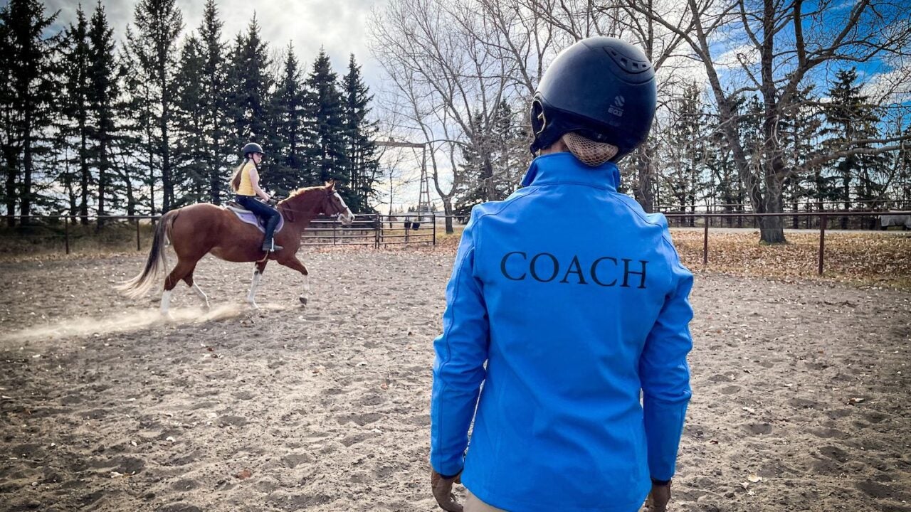 Coach in a blue jacket standing in an arena giving a lesson to a young lady on a chestnut horse