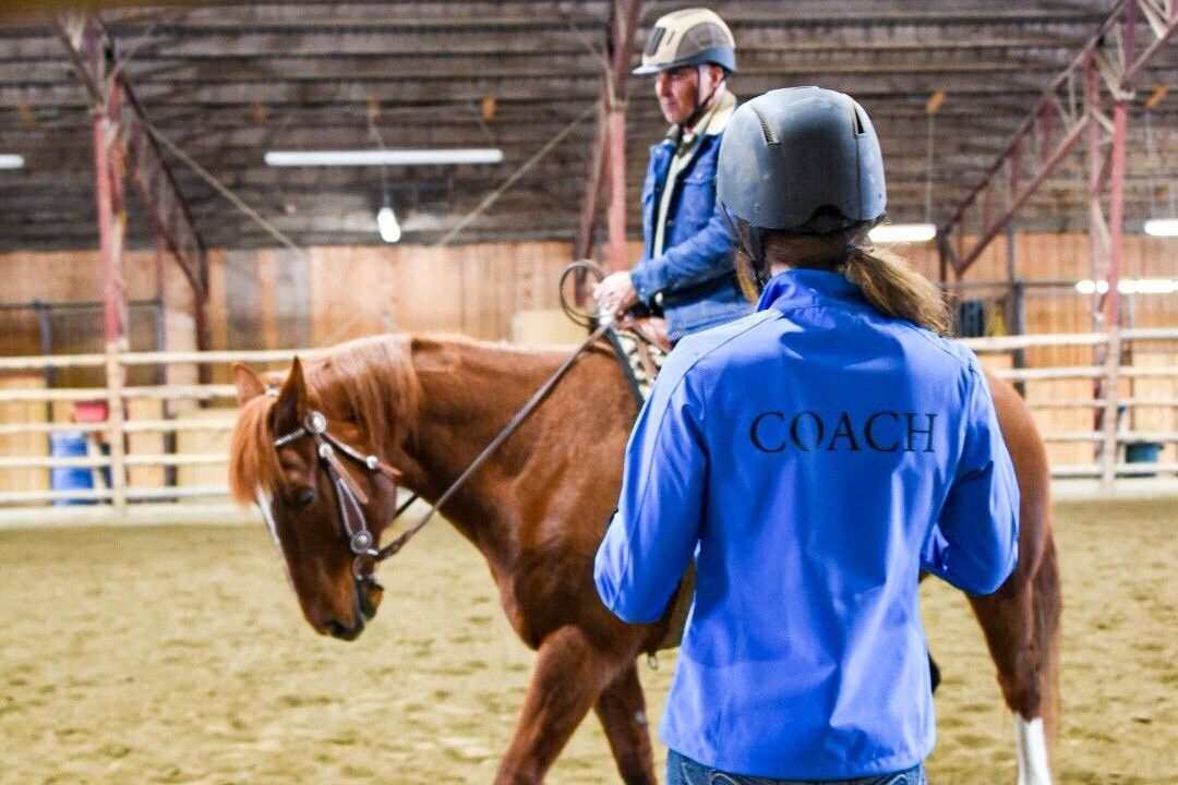 Coach with a blue jacket teaching a male riding western and wearing a helmet