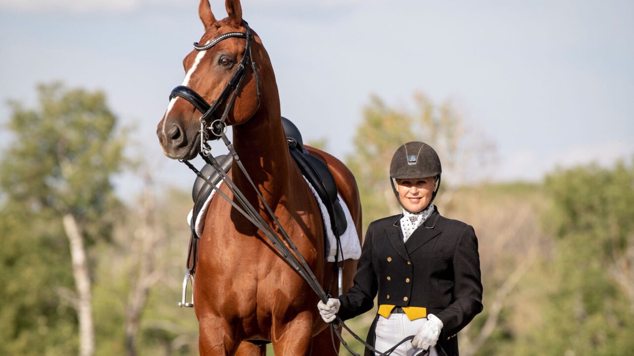 Lady in a black dressage tailcoat and black helmet standing beside a tall chestnut horse