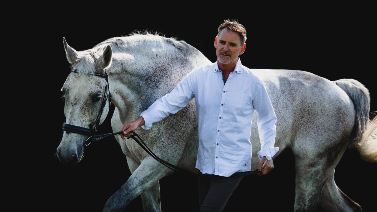 Man in white shirt leading a tall flea bitten grey horse in a bridle